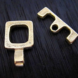 Pode incluir: Um fecho de metal dourado com um laço quadrado e uma barra retangular com um furo no centro.