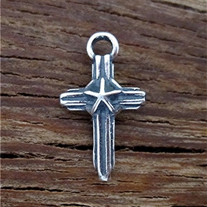 May include: A silver charm in the shape of a cross with a five-pointed star in the center. The cross has a textured, ridged design.