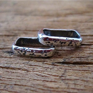 May include: Two silver-colored, rectangular rings with a hammered texture. The rings are placed on a wooden surface. The rings have a hollow center and rounded edges. The metal has a slightly reflective surface.