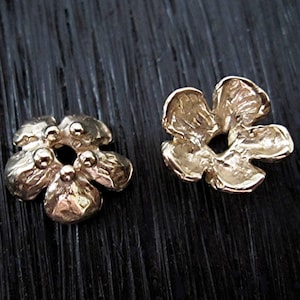 May include: Two gold-colored flower-shaped jewelry components. Each component has five petals and a central hole. One has small, round accents. The components are likely for jewelry making or embellishment.
