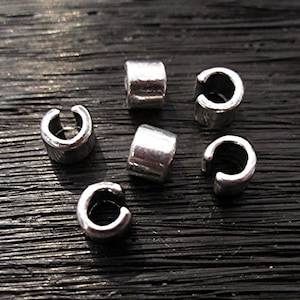 May include: Close-up of six small, silver-colored metal ear cuffs. The ear cuffs are cylindrical with a C-shaped opening. The background is a dark, textured surface.