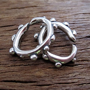 May include: Two silver-colored hoop earrings with a textured design. Each earring has a series of small, round embellishments along the outer curve. The earrings are displayed on a rustic, wooden surface.