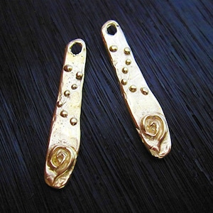 Op de afbeelding: Twee goudkleurige metalen bedels met een getextureerd oppervlak en een reliëf hartontwerp aan de onderkant. De bedels hebben een kleine lus aan de bovenkant om aan sieraden te bevestigen.