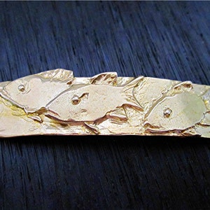May include: A gold-colored metal bar featuring three stylized fish. The rectangular bar has rounded ends with a loop on each end. The fish are depicted in relief, with details like fins and eyes.