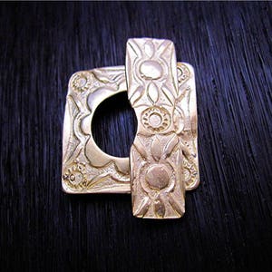 May include: A gold-toned metal buckle with an intricate design. The buckle is square with a round opening in the center. The design features a repeating pattern of stylized flowers and leaves.
