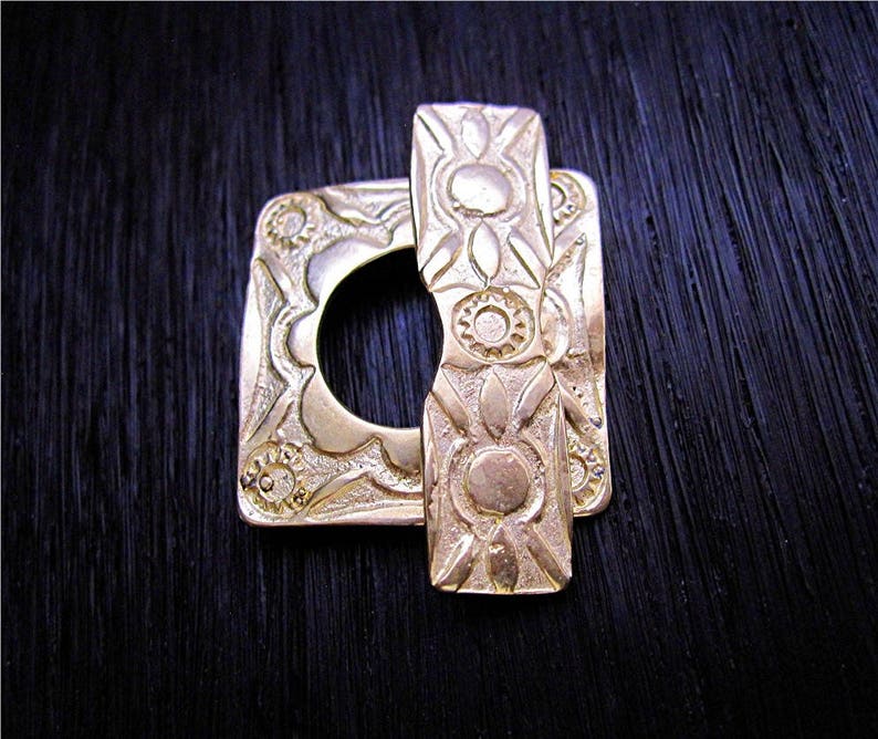 May include: A gold-toned metal buckle with an intricate design. The buckle is square with a round opening in the center. The design features a repeating pattern of stylized flowers and leaves.