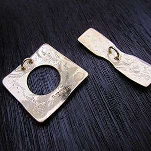 May include: Two gold-toned metal buttons with a textured surface. One button is square with a round hole in the center. The other button is a long oval shape.
