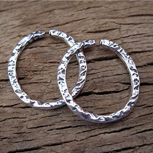 May include: A pair of silver hoop earrings featuring a textured, hammered surface. Each earring is circular with a small opening at the top, showcasing a reflective metallic finish with deep indentations across the entire outer circumference.