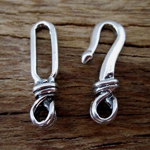 May include: Two silver-colored metal clasps. One is a hook shape, and the other is a loop. Both clasps have a decorative twisted design at the base, connecting the hook and loop elements. The background is a wooden surface.