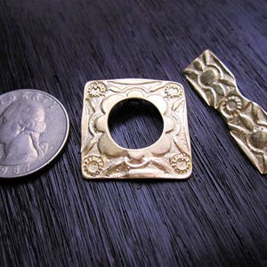 May include: Two gold-toned metal decorative pieces. One is a square with a circular cutout and intricate floral designs. The other is a long, narrow piece with a bow-like shape and floral designs.