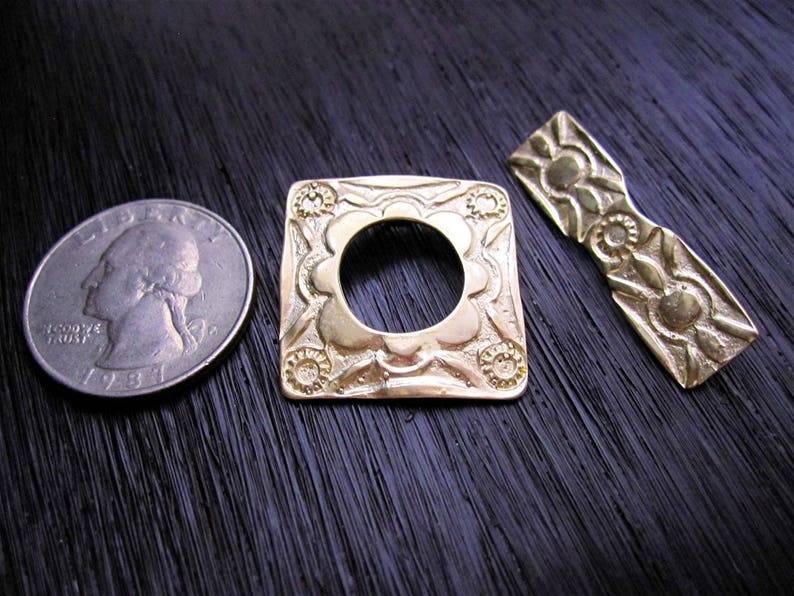 May include: Two gold-toned metal decorative pieces. One is a square with a circular cutout and intricate floral designs. The other is a long, narrow piece with a bow-like shape and floral designs.