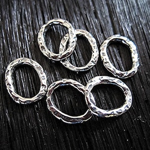 Op de afbeelding: Zes zilverkleurige ovale sieraden schakels met een gehamerde textuur. De schakels liggen op een donkere, getextureerde ondergrond. De schakels hebben een rustieke, handgemaakte uitstraling.