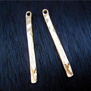 May include: Two gold-colored, hammered metal bar pendants with a small hole at the top for stringing. The bars have a slightly irregular shape and a textured surface. These pendants are suitable for jewelry making.