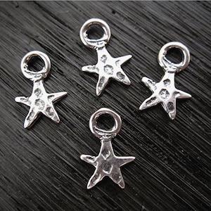 May include: Four small, silver-colored starfish charms with textured surfaces. Each charm has a loop at the top for attaching to jewelry. The charms are arranged on a dark, textured surface.