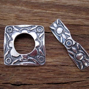 May include: Two silver metal decorative pieces with intricate floral designs. The piece on the left is square with a circular opening in the center. The piece on the right is a long, narrow rectangle.