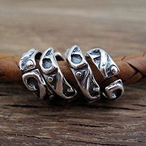 May include: A set of five silver-colored metal beads with an abstract design. The beads are arranged in a spiral on a brown leather cord, set against a wooden background. The beads have a textured, organic appearance.