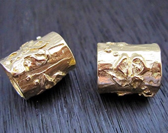 May include: Two gold-colored, cylindrical rings with a textured surface and floral design. The rings are displayed on a dark wooden surface. The rings have a rustic, organic aesthetic.