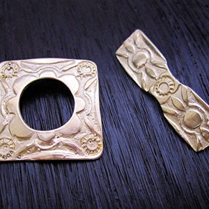 May include: Two gold-toned metal decorative pieces with intricate floral designs. One piece is a square with a circular opening in the center, the other is a long, narrow rectangle.