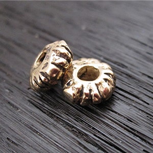 May include: Two gold-colored metal beads with a textured, ridged design. The beads have a circular shape with a hole in the center.