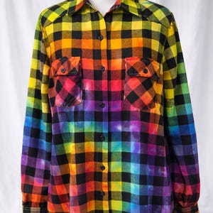 May include: Rainbow plaid button-down shirt with two chest pockets. The shirt features a vibrant gradient of colors, including yellow, orange, red, purple, and blue, transitioning into a black and white checkered pattern. The long-sleeved shirt is displayed on a white mannequin.