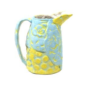 Può includere: Brocca in ceramica fatta a mano con corpo azzurro e beccuccio e base gialli. La brocca presenta un motivo floreale sulla parte superiore e un motivo a pois sulla parte inferiore. Il manico è azzurro.