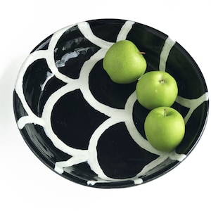 May include: A black ceramic bowl with a white geometric pattern, filled with three green apples.
