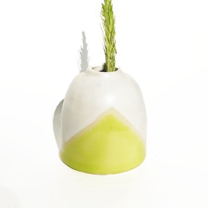 Könnte beinhalten: Eine kleine, weiße Keramikvase mit einem grünen geometrischen Muster. Ein einziger grüner Grashalm steht in der Vase.