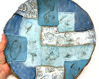 May include: A handcrafted ceramic plate with a patchwork design. The plate features various shades of blue and white, with textured patterns including polka dots and leaf imprints. The plate is round with a slightly irregular edge.