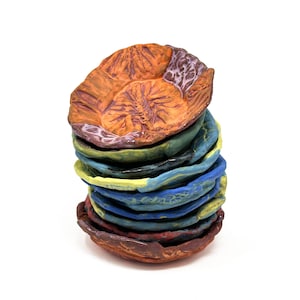 Puede incluir: Una pila de platos de cerámica hechos a mano en varios colores. El plato superior es naranja con una superficie texturizada y un patrón detallado. Otros platos son azules, verdes y amarillos. Los platos están apilados uno encima del otro.