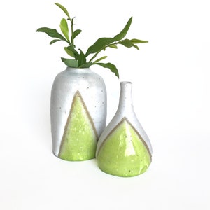 Könnte beinhalten: Zwei Keramikvasen mit weißer Glasur und einem grünen geometrischen Design. Die höhere Vase hat eine zylindrische Form und die niedrigere Vase eine abgerundete Form. Beide Vasen sind mit grünen Blättern gefüllt.