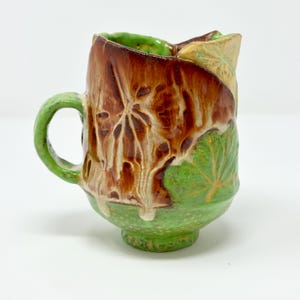 Op de afbeelding: Een groene en bruine keramische mok met een bladerenontwerp. De mok heeft een handvat en een afgeronde basis.