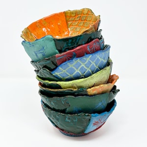 Puede incluir: Una pila de cuencos de cerámica hechos a mano en varios colores, incluyendo naranja, turquesa, rojo, azul y verde. Cada cuenco tiene un diseño y borde texturizado único. Los cuencos están apilados uno encima del otro, creando una exhibición visualmente atractiva.