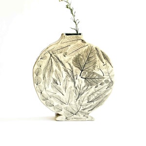 May include: A white ceramic vase with a round body and a wide opening. The vase is decorated with a pattern of pressed leaves in black.