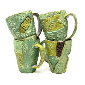 Pode incluir: Quatro canecas de cerâmica empilhadas em tons de verde e amarelo. Cada caneca apresenta um padrão único de folhas e flores em relevo. As alças são curvas e as canecas têm uma aparência rústica e artesanal.