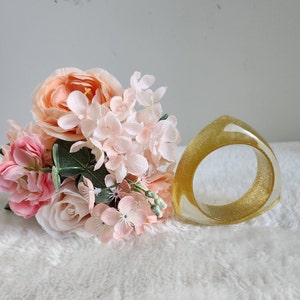 May include: A gold-colored resin ring with a triangular shape and a smooth, glossy finish. The ring is displayed on a white surface with a bouquet of pink and peach flowers.