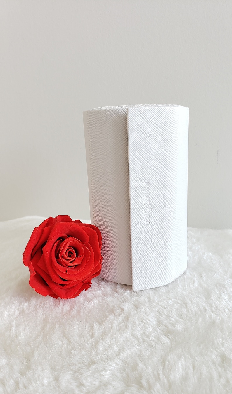 May include: A white cylindrical box with the word "Pandora" embossed on the side. The box is sitting on a white fluffy surface next to a single red rose.