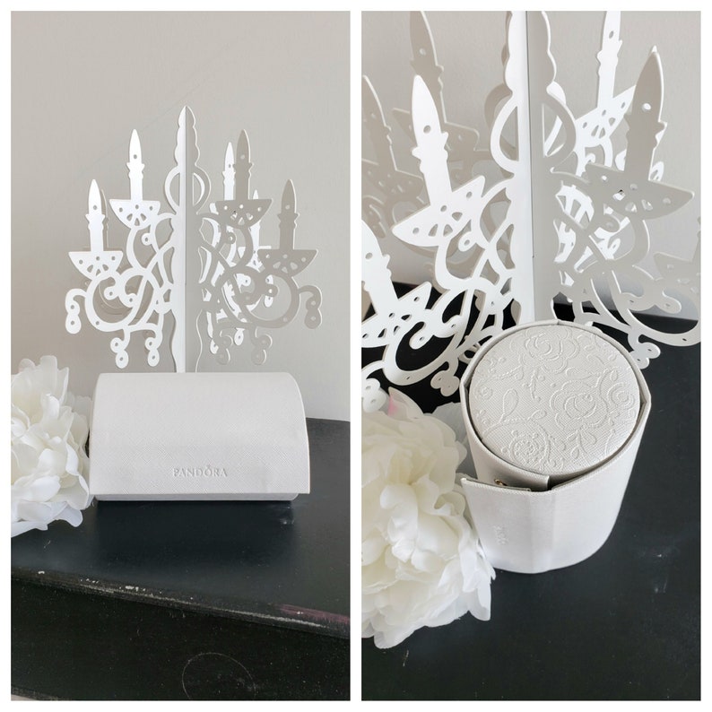 May include: White jewelry box with a decorative, ornate, chandelier-shaped lid. The box is made of cardboard and has a white satin interior. The box is labeled "Pandora" on the front.