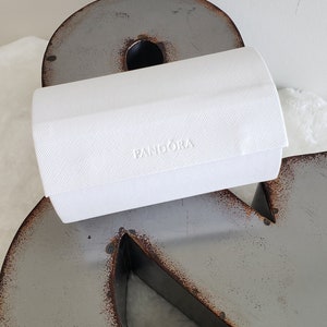 May include: A white rectangular box with the word "PANDORA" embossed on the top. The box is sitting on a gray metal surface.