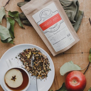 May include: A brown paper bag with the text "Winterwoods Tea Company" and "Harvest Apple" printed on it. The bag contains loose leaf tea and has a recommended brewing guide. A white plate with a cup of tea and a slice of apple in it. A red apple on a green branch.