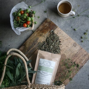 May include: A brown paper bag with a white label that reads "Farmers Market Mint" with a green leaf design. The bag is filled with loose leaf tea and is sitting on a wooden cutting board. A cup of tea and a basket of tomatoes are also in the image.