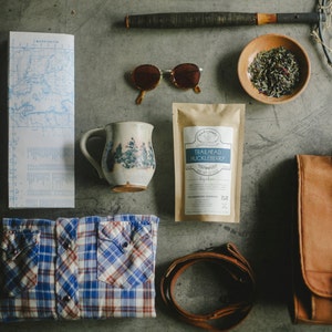 May include: A flat lay of various items including a map, a mug with a floral design, a bag, sunglasses, a tea bag, a bowl of tea leaves, and a leather strap.