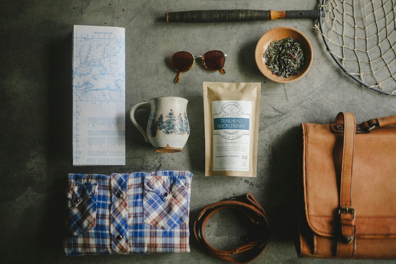 May include: A flat lay of various items including a map, a mug with a floral design, a bag, sunglasses, a tea bag, a bowl of tea leaves, and a leather strap.