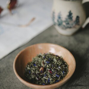 May include: A wooden bowl filled with loose leaf tea. The tea is a blend of green tea, dried flowers, and herbs.