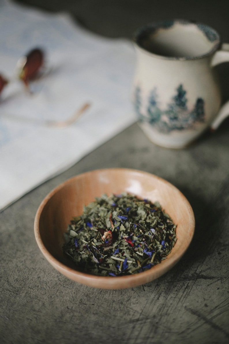 May include: A wooden bowl filled with loose leaf tea. The tea is a blend of green tea, dried flowers, and herbs.