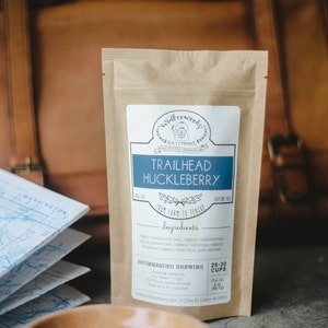 May include: A brown paper bag with a label that reads "Winterwood Tea Company" and "Trailhead Huckleberry". The label also includes the ingredients and recommended brewing instructions. The bag is sitting on a table with a wooden bowl of loose leaf tea.