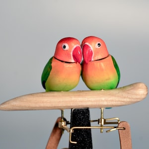 Peut inclure: Deux inséparables en bois perchés sur une branche marron clair. Les oiseaux ont la tête rouge, le corps orange, les ailes vertes et un bec rose vif. Les oiseaux font partie d'une pièce décorative avec une base noire et des accents métalliques dorés.