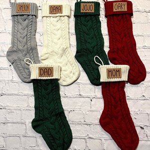 May include: Six cable-knit Christmas stockings in various colors: gray, cream, green, and red. Each stocking has a cream-colored cuff and a brown leatherette tag with a name: Cameron, Dana, Jojo, Gary, Dad, and Mom. A loop is attached for hanging.