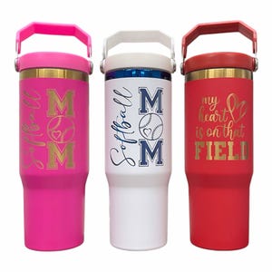 May include: Three insulated tumblers with handles. One is pink with "Softball MOM" in gold lettering, another is white with "Softball MOM" in blue, and the third is red with "my heart is on that FIELD" in gold.