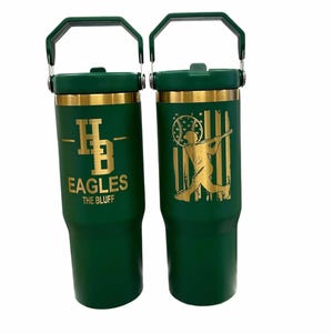 May include: Two green insulated tumblers with gold accents and handles. One features "HB EAGLES THE BLUFF" in gold lettering. The other has a gold baseball player silhouette against a gold and white flag design.