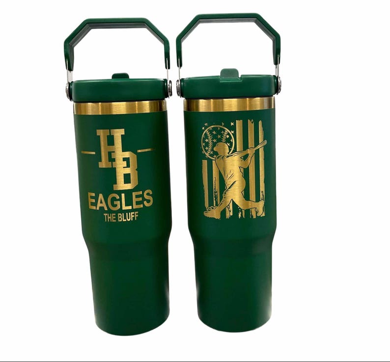 May include: Two green insulated tumblers with gold accents and handles. One features "HB EAGLES THE BLUFF" in gold lettering. The other has a gold baseball player silhouette against a gold and white flag design.